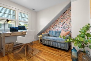 A room with a desk, chair, and a sofa. There is a window with blinds, a patterned wall, and a plant.