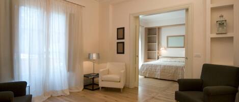 A hotel room with a wooden floor, a bed, a white armchair, a dark armchair, a small table with a lamp, and a door leading to another room.