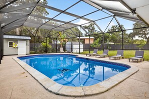 Ein überdachter Pool im Freien mit blau gefliestem Rand, umgeben von Liegestühlen und einer kleinen Gartenhütte.