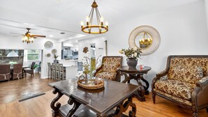 A spacious living room with a wooden coffee table, patterned armchairs, a chandelier, and a kitchen in the background.