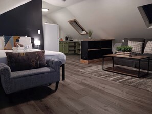 Een moderne slaapkamer in loftstijl met een bed, een grijze fauteuil, een bijzettafeltje en een inbouwkast voor de televisie.