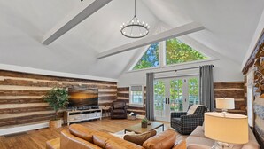 Ein großzügiger Wohnraum mit hoher Decke, Holzwänden, einem großen Fernseher, Ledersofas und einem Kronleuchter.