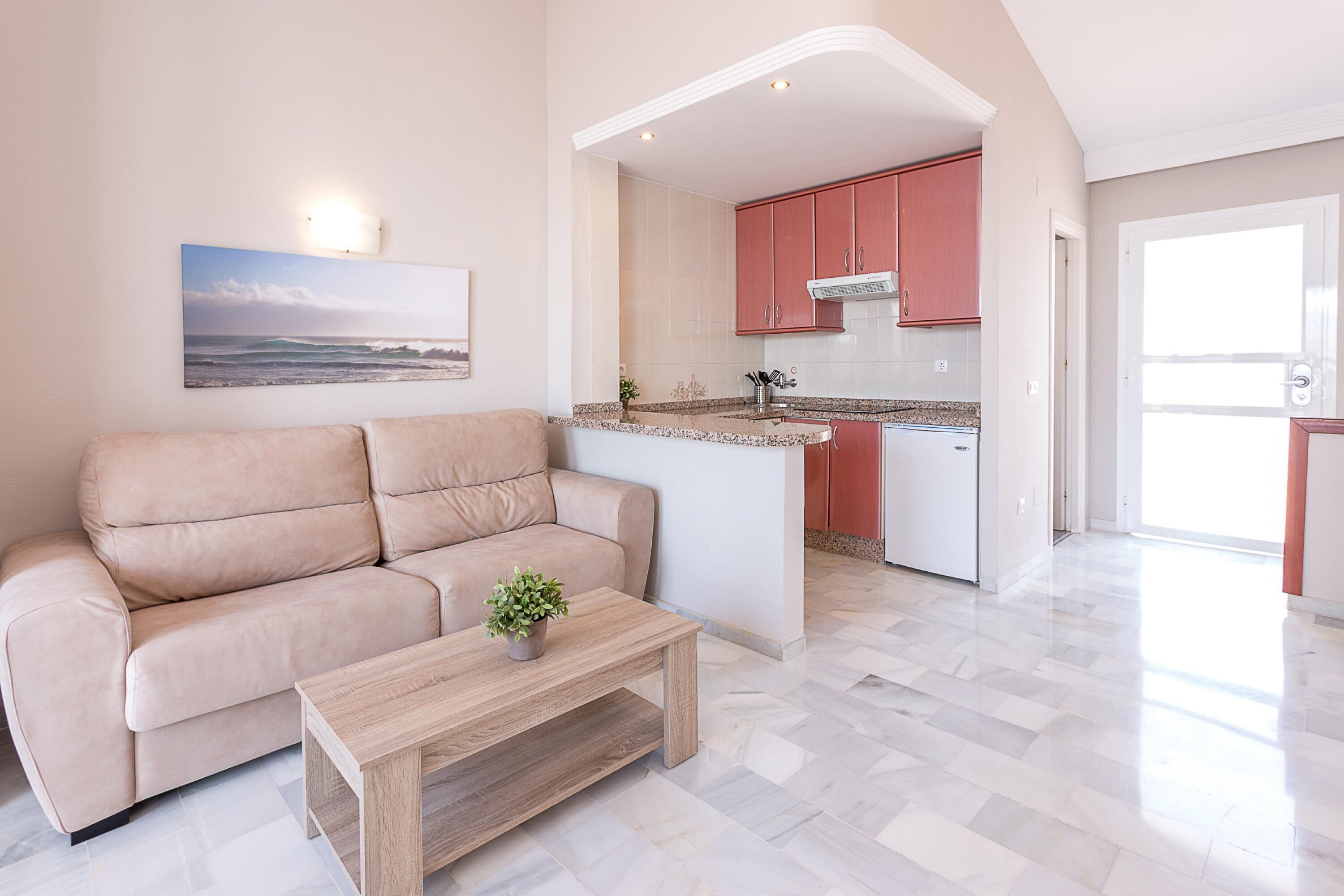 Un salon moderne avec un canapé beige, une table basse en bois et un coin cuisine doté de meubles rouges et d’un îlot central blanc.