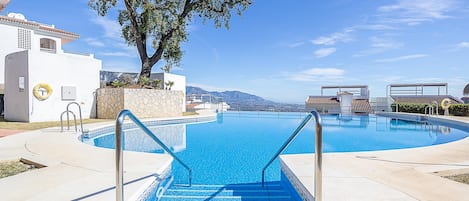 Une piscine avec des marches permettant d’y accéder, entourée d’un bâtiment blanc et d’un grand arbre.