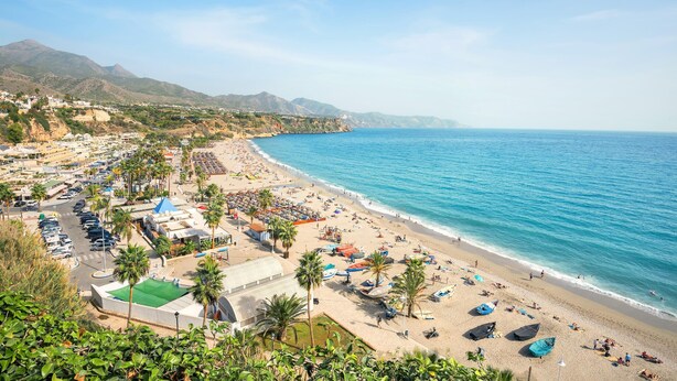 A coastal beach scene with a clear view of the sea, a sandy beach, and a parking area with cars and a green-roofed building. - Nerja City Center Apartment (Nerja)