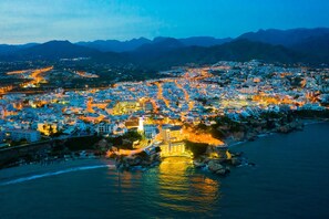 Une vue d’ensemble d’une ville côtière au crépuscule, avec des bâtiments illuminés, un phare imposant et une chaîne de montagnes en arrière-plan.