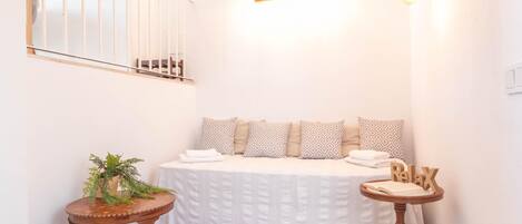 Un dormitorio con cama, una mesita con una planta, una mesita de noche con un libro, y una lámpara fijada a la pared.