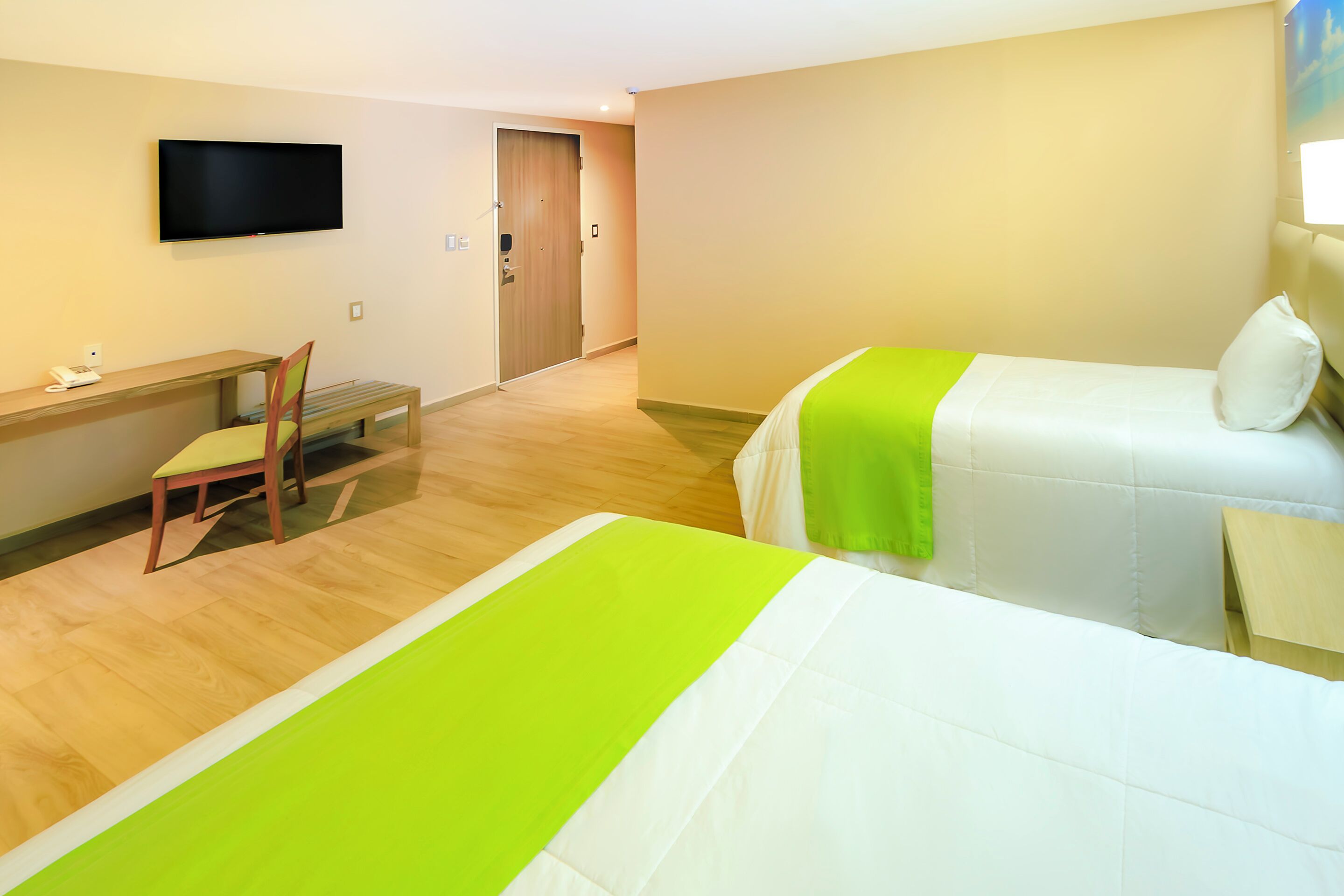 Quarto de hotel com duas camas, uma escrivaninha, uma cadeira, uma televisão e uma porta.