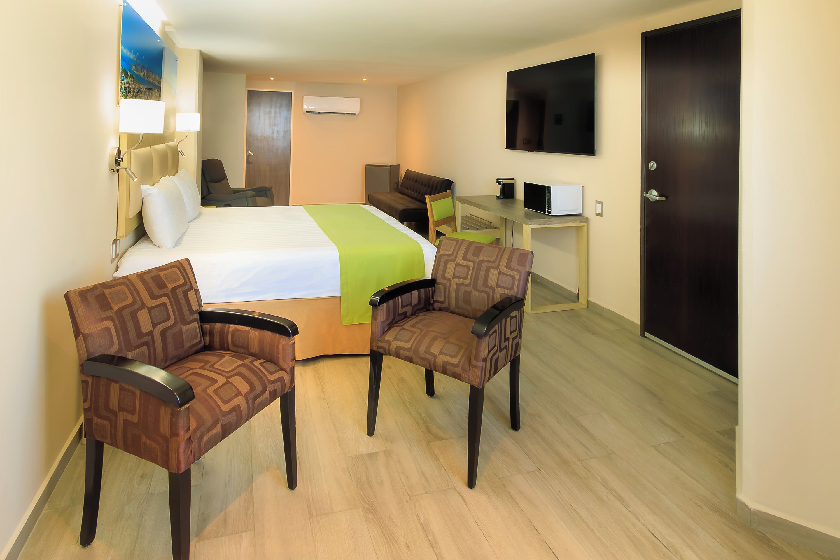 A hotel room with a bed, two chairs, a desk, a TV, and a door.