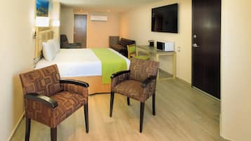 A hotel room with a bed, two chairs, a desk, a TV, and a door.