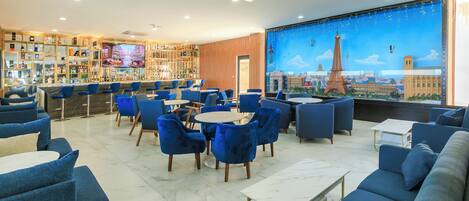 Un rincón de bar con sillas y mesas azules, un televisor y un mural de la Torre Eiffel.