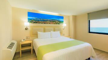 Une chambre d’hôtel avec un lit, une table de chevet, une lampe, un tableau représentant un paysage urbain côtier et une fenêtre donnant sur la mer.