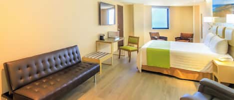 Una habitación de hotel moderna con una cama grande, un sofá de cuero, un escritorio con una silla y un ventanal que proporciona luz natural.