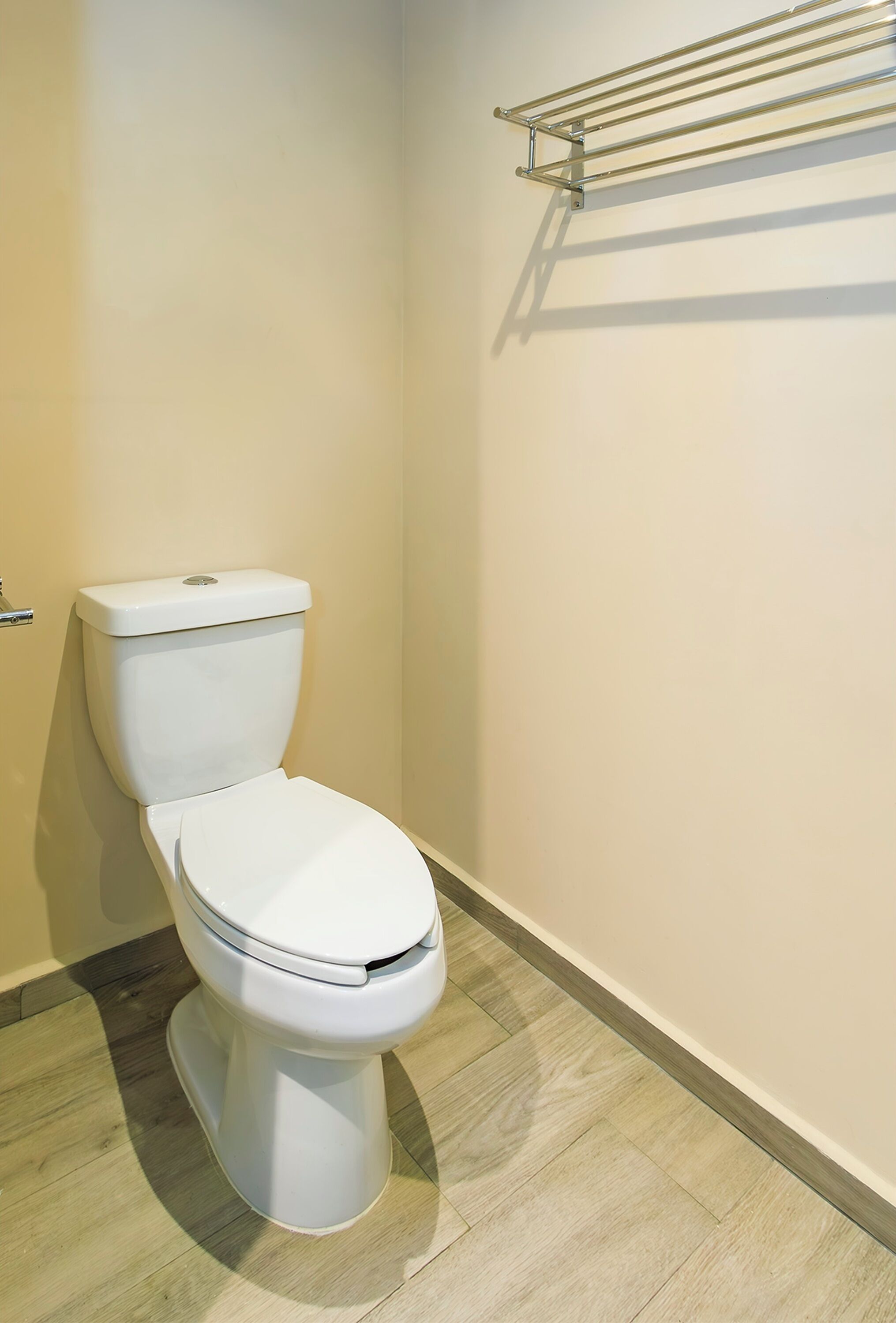 A white toilet with a metal rack above it, a wooden floor, and a plain wall.