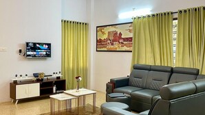 A modern living room with a sectional sofa, a flat-screen TV, a small coffee table, and a decorative painting on the wall. - Casa Grey Trivandrum (Thiruvananthapuram)