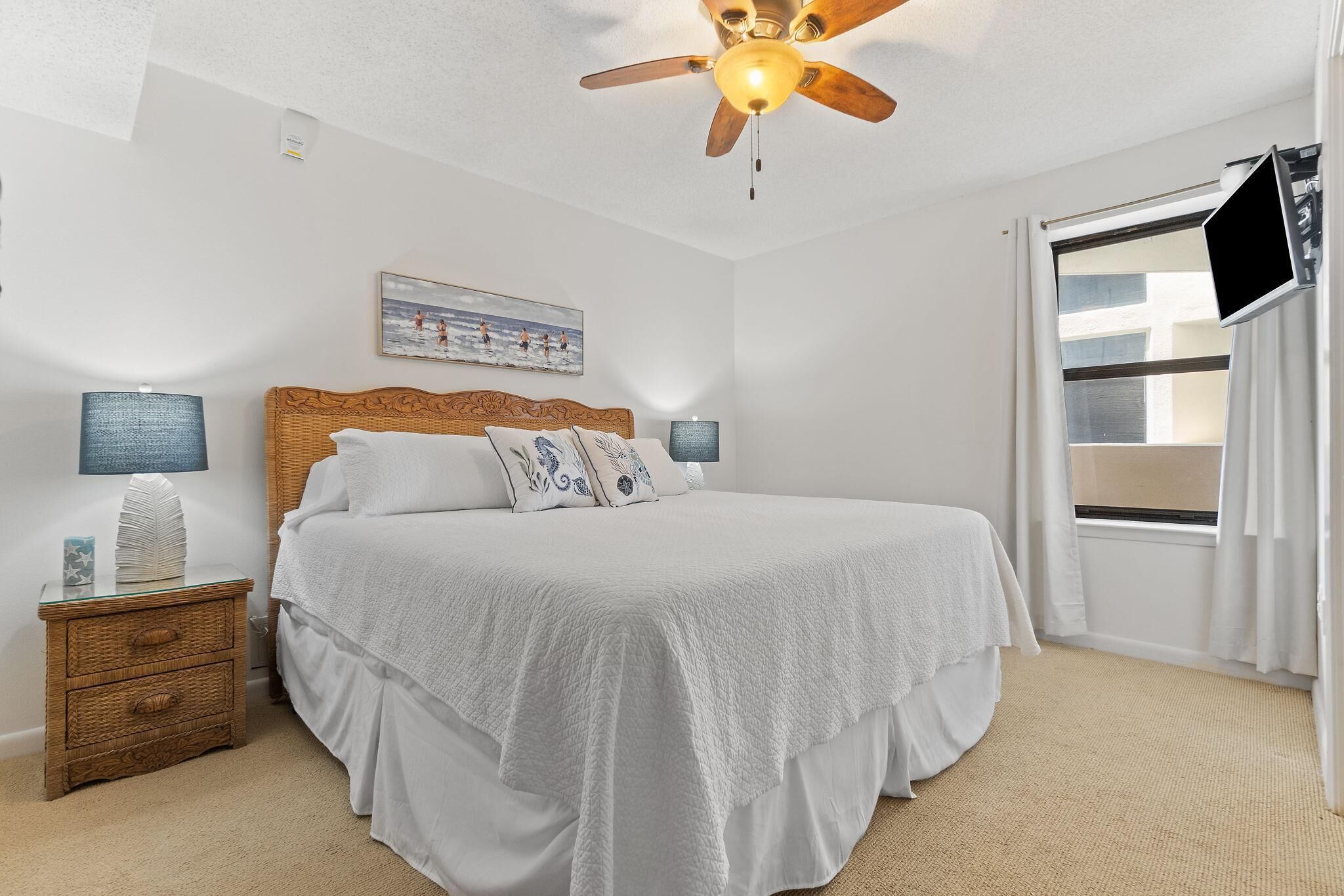 A bedroom with a bed, a ceiling fan, a nightstand, a lamp, a painting, and a window with curtains.