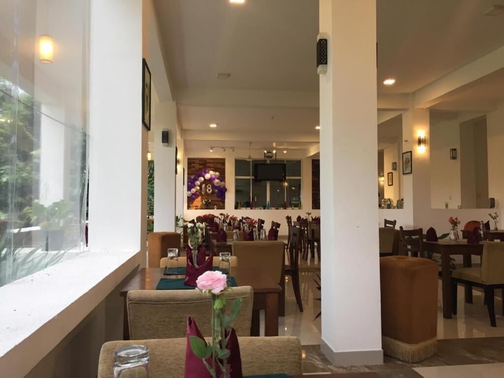 Um hall de hotel espaçoso, com várias áreas de refeições, uma grande janela com vista para vegetação e um arranjo floral decorativo central.