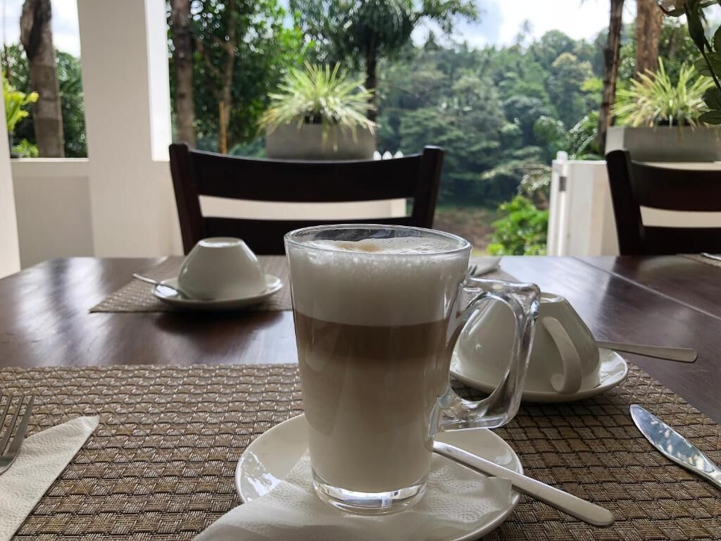 A table with a glass of coffee, a mug, and a saucer, set on a woven placemat.