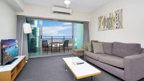 A modern living room with a grey sofa, a coffee table, a TV on a stand, and a balcony with a view of the sea. - Pandanas Apt 7 (15th fl with harbour & city views) (Darwin City)