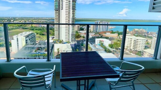 A balcony with a table and chairs, overlooking a cityscape with tall buildings and a clear sky. - Pandanas Apt 8 (18th fl, Sunrise & Harbour Views) (Darwin City)