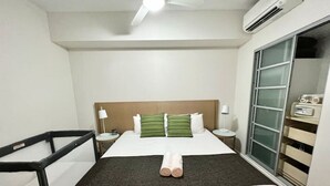 A hotel room with a bed, a desk, a chair, a wardrobe, and a ceiling fan. - Pandanas Apt 8 (18th fl, Sunrise & Harbour Views) (Darwin City)