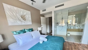 A hotel room with a bed, a nightstand, a wall-mounted artwork, a bathroom with a glass shower enclosure, and a wooden floor.