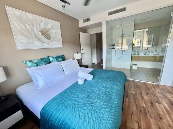 A hotel room with a bed, a nightstand, a wall-mounted artwork, a bathroom with a glass shower enclosure, and a wooden floor. - Waterfront Escape (2 bedrooms) (Darwin City)