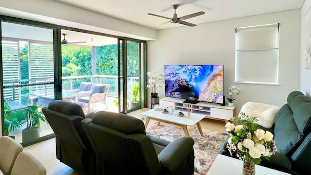 A modern living room with a flat-screen TV, comfortable seating, a ceiling fan, and a balcony with outdoor furniture.