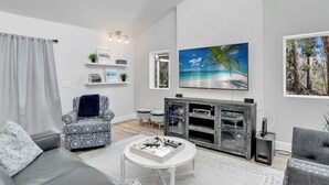 A modern living room with a grey sofa, patterned armchair, and ottoman, a white coffee table, and a fireplace with a TV above it.