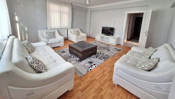Een moderne woonkamer met twee witte banken, een centraal ottoman, een flatscreen-televisie en een kamerkleed met een patroon.