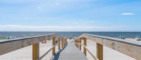 En træpromenade fører til en sandstrand med parasoller og strandstole.