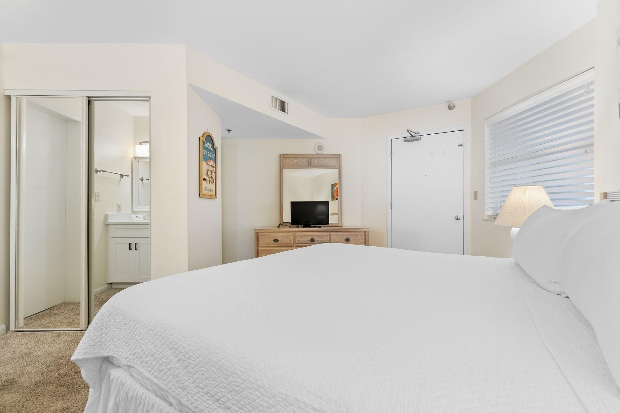 Un dormitorio con una cama grande, un televisor sobre un aparador de madera, un espejo y una puerta que da a un baño.