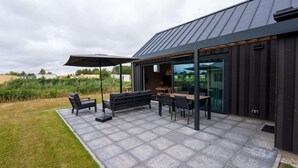 A modern outdoor patio with a table and chairs, a large umbrella, and a glass door leading to a covered area. - Villa in Breskens Near the Beach (Breskens)