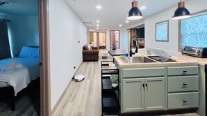 Ein modernes Hotelzimmer mit Bett, einer Sitzecke, einer Küchenzeile inklusive Spüle und Toaster sowie einem Flur, der zu einem weiteren Zimmer führt.
