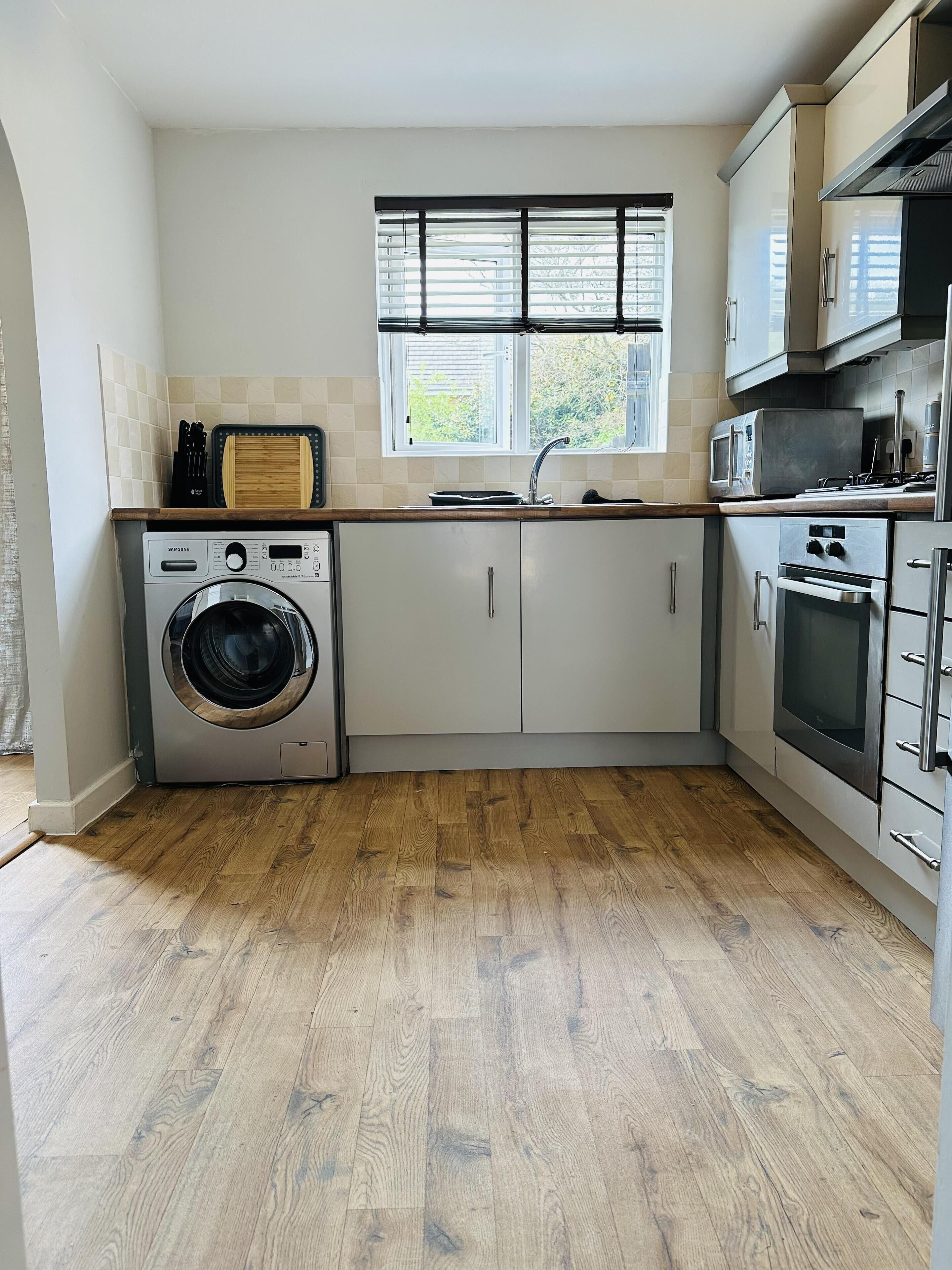 A compact kitchen with a washing machine, microwave, oven, and a window with blinds.