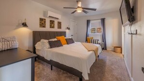 Quarto com cama, mesas de cabeceira, ventoinha no teto, televisão na parede, obra de arte e janela com cortinas.