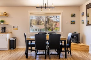 A dining area with a wooden table and chairs, a window with a view of trees and houses, a black stove, and a chandelier. - Cozy Central Forest Retreat (Casper)