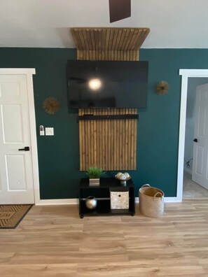A room with a wooden panel wall, a mounted flat-screen TV, a small black console table with decorative items, and a woven basket.