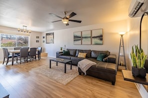 A modern living room with a dining area, a ceiling fan, a large window, and a potted plant.