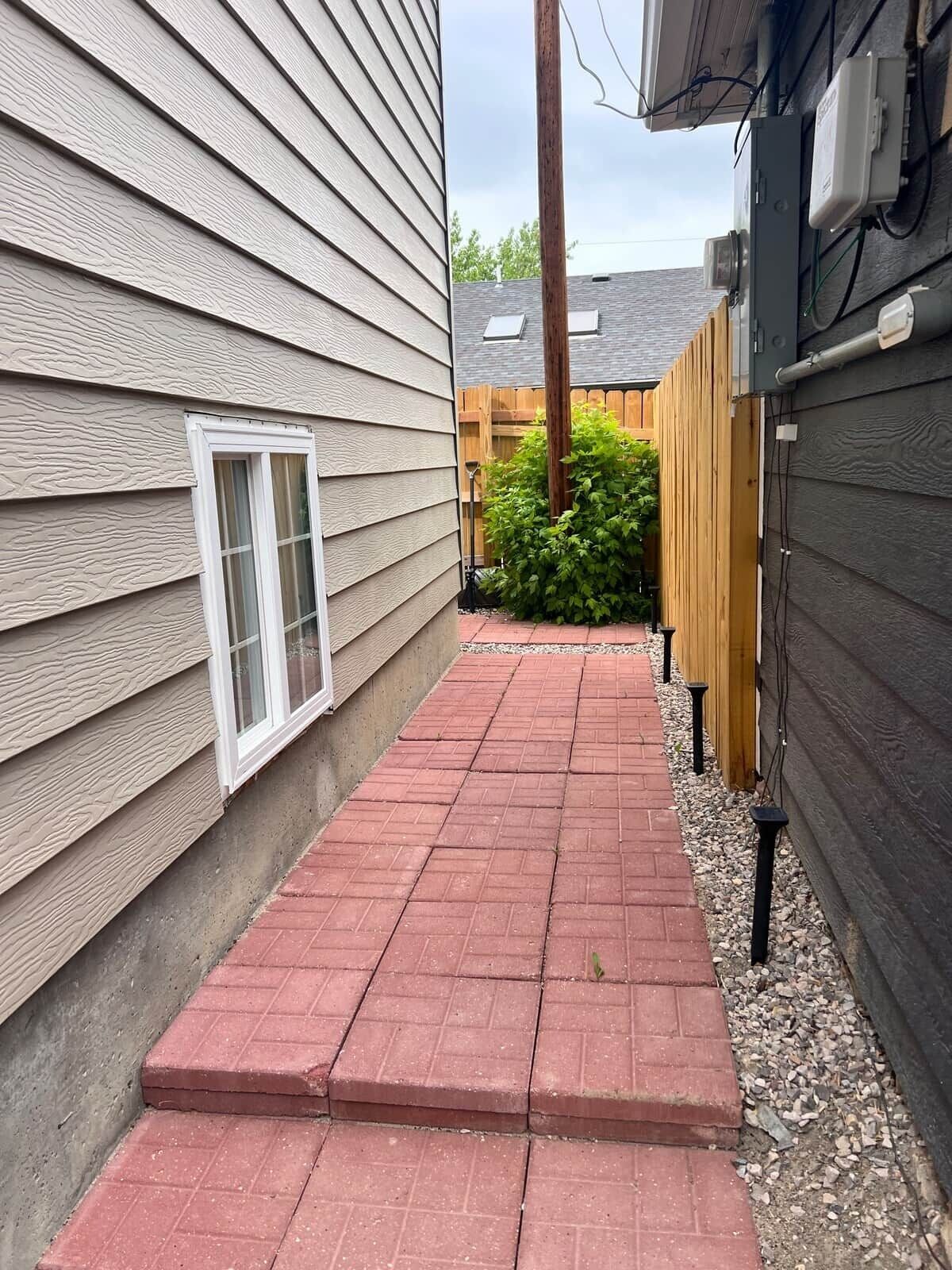 A narrow brick pathway leading to a wooden fence, with a window on the left and a utility box on the right.