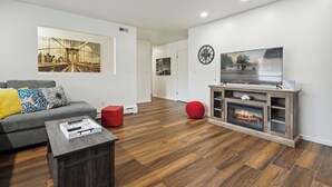 A modern living room with a grey sofa, a red ottoman, a wooden coffee table, a fireplace, and a large TV on a wooden stand.
