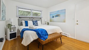 A bedroom with a bed, a bench, a window with blinds, a painting, and a door.