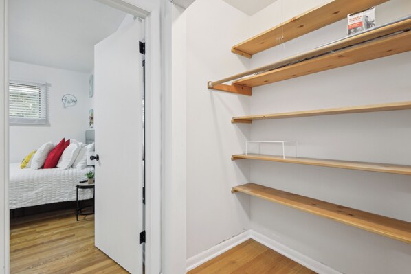 A bedroom with a bed, a nightstand, and a window with blinds. There are wooden shelves against the wall.