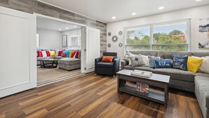 Un salón amplio con un sofá gris, un sillón negro y una mesa de centro. Hay una puerta que da a otra habitación, y una ventana con persianas.