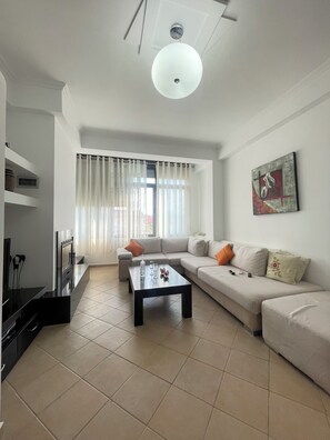 A modern living room with a sofa, a coffee table, and a TV. There is a window with curtains and a painting on the wall. - Apartment in private house (Tirana)
