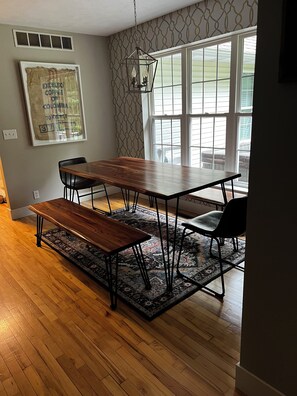 Een eetruimte met een houten tafel en bankjes, behang met een patroon en grote ramen.