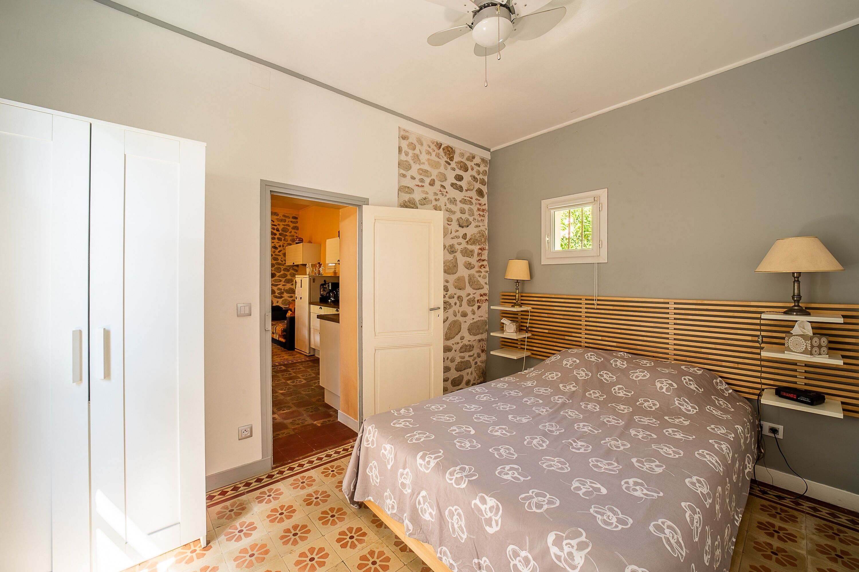 A bedroom with a bed, a wardrobe, a stone wall, a window, and a ceiling fan.