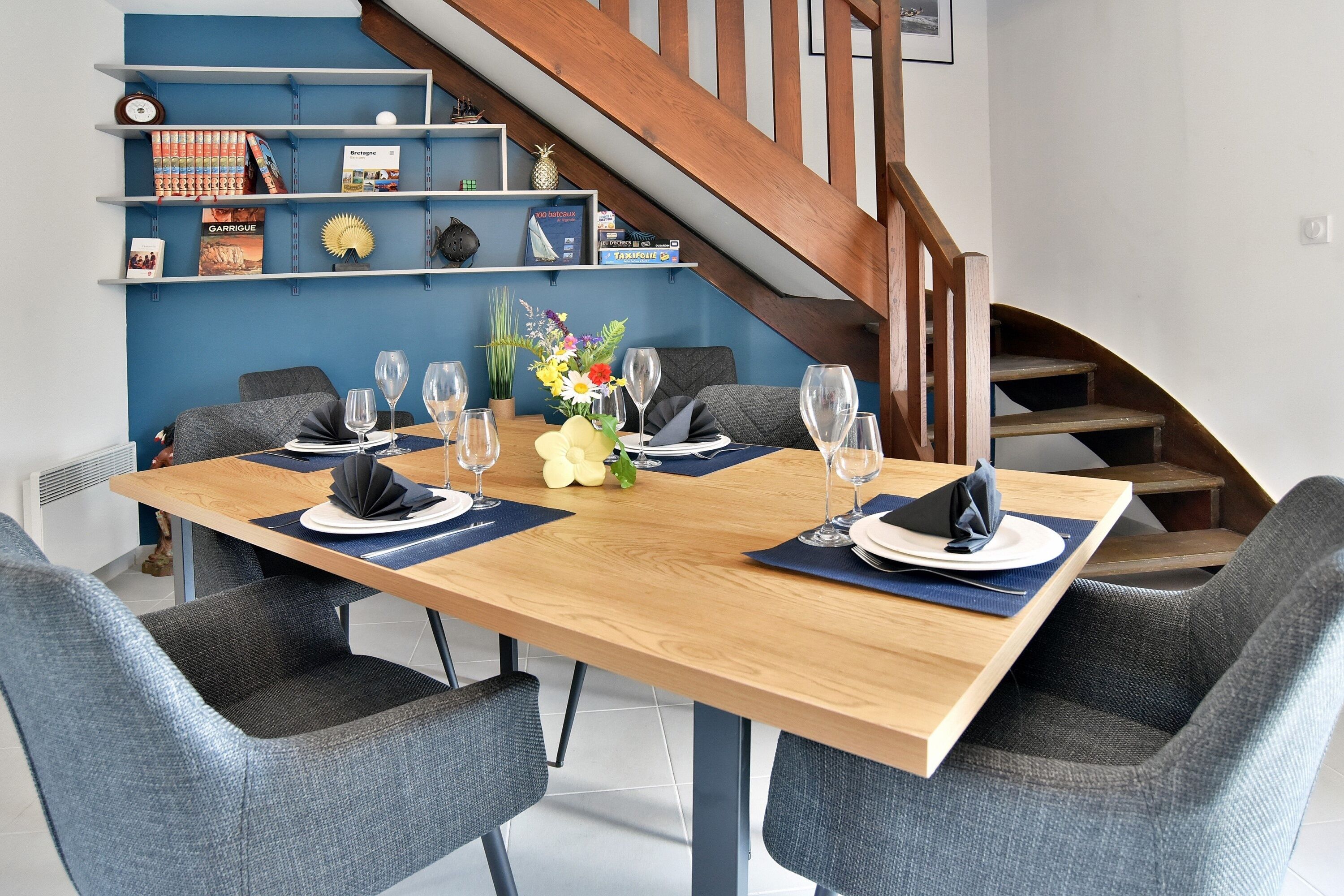 Un coin repas avec une table en bois dressée pour quatre personnes, entourée de chaises à revêtement gris. Un escalier avec des rampes en bois est visible à l’arrière-plan.