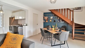 A modern living room with a dining area, a kitchen in the background, and a wooden staircase. - Duplex Plancoët - Pour 4p, à 15 min de la plage (Créhen)