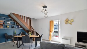 A modern living room with a staircase, dining area, and a TV on a stand. - Duplex Plancoët - Pour 4p, à 15 min de la plage (Créhen)